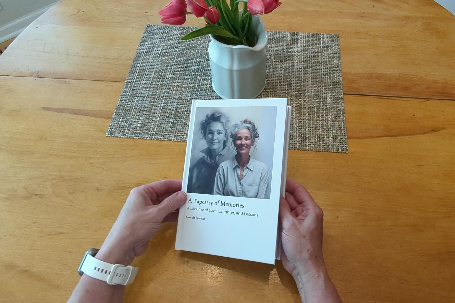 A Kindred Tales hardcover memoir, A Tapestry of Memories, held in two hands on a kitchen table beside a vase of tulips